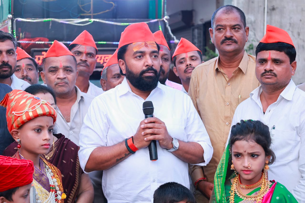Rahul Awade speaking into a microphone at a street programme in Ichalkaranji, wearing a saffron pheta, with children in traditional Maharashtrian dress beside him.